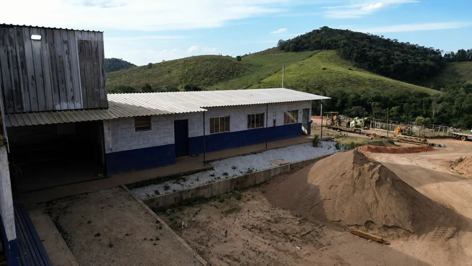Galpão e prédio de alvenaria em área de mineração, com serras cobertas de vegetação ao fundo